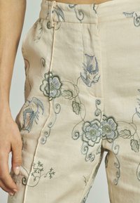 Beige broek met blauwe en groene bloemen- en vogelborduring, met een hoge taille en een zijzak, gedragen door een persoon met een zichtbare hand.