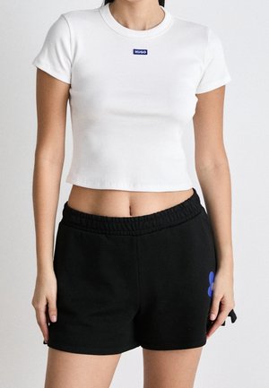 Kvinde iført en hvid cropped T-shirt med "HUGO" logo og sorte shorts med elastisk talje og et lille blåt blomstermotiv på siden.