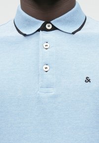 Gros plan sur un polo bleu clair avec un bord noir sur le col, trois boutons blancs et un petit symbole esperluette noir brodé sur la poitrine.