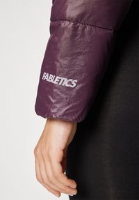 Manica di giacca a vento viola con orlo cucito e logo "FABLETICS" bianco. Tessuto liscio e lucido con polsino elasticizzato.
