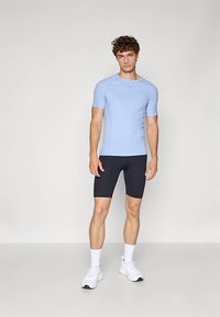 Fiatal férfi áll, világoskék, testhez simuló rövid ujjú ingben, fekete kerékpáros rövidnadrágban, fehér zokniban és fehér sportcipőben, egyszínű háttér előtt.