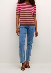 Persona vestida con una blusa corta de rayas rosa y marrón, jeans de mezclilla azul claro y sandalias de tacón marrón, de pie contra una pared sencilla.