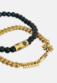 Melna pērlu rokassprādze ar zelta ķēdi un akcents, kurai ir ziedu raksts uz zelta pērlītes un birka ar izcirstu uzrakstu "ICON".