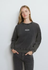 Sudadera gris de gran tamaño con cuello redondeado acanalado y mangas desgastadas. Presenta un parche de logo blanco en el centro del frente.