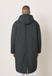 Muž oblečený v dlhom čiernom kapucňovom kabáte stojaci chrbtom ku kamere na jednoduchom svetlom pozadí.