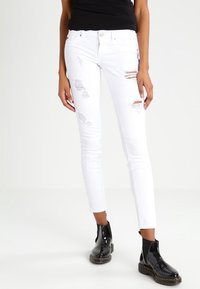 Jean skinny blanc avec détails usés et patches effilochés. Associé à des bottines noires brillantes avec une semelle épaisse et une fermeture éclair latérale.
