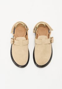 Beige semskede sandaler med en bred justerbar rem og spenne-detalj. Svart gummisåle med gul søm rundt kanten for kontrast.
