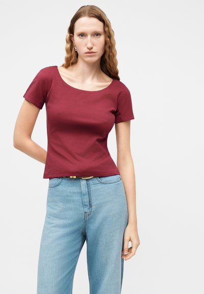 Γυναίκα με μακριά κυματιστά μαλλιά που φοράει ασημένια σκουλαρίκια, στενό μπορντό μπλουζάκι με κοντά μανίκια και ψηλόμεσα ανοιχτόχρωμα μπλε τζιν, μπροστά σε λευκό φόντο.