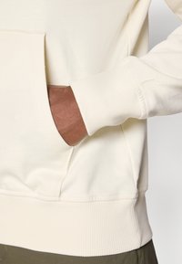 Kremowy sweatshirt z przednią kieszenią, ściągaczami na rękawach i dolnej krawędzi. Miękka tkanina i luźny krój, zaprezentowany w połączeniu z oliwkowymi spodniami.