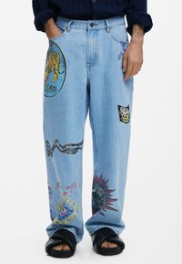 Hellblaue Jeans mit weitgeschnittenen Beinen, die verschiedene bunte Grafikdrucke aufweisen, darunter einen Tiger, eine Sonne und abstrakte Designs auf den Taschen.