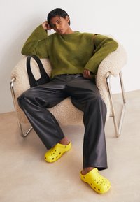 Green textured oversized sweater, black leather wide-leg pants, and bright yellow clogs with ventilation holes, seated on a fluffy chair.