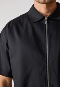 Μαύρο κοντομάνικο πουκάμισο με φερμουάρ μπροστά. Διαθέτει κλασικό γιακά και απαλό, υφασματώδες ύφασμα. Το ασημένιο φερμουάρ προσθέτει αντίθεση.