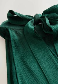 Groene stof met textuur, afgewerkt met een strikdetail en vloeiende banden. Het materiaal lijkt lichtgewicht en licht glanzend.