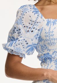 Valge puff-sleeve kleit siniste lillede broderi ja lõigatud detailidega. Rikkalikult kokku volditud bodice ning volangidega varrukate ääred toovad disaini esile.