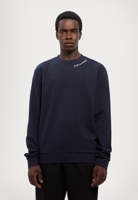 Granatowy sweatshirt z okrągłym dekoltem, długimi rękawami i prążkowanymi mankietami. Z przodu, na lewym ramieniu, znajduje się małe białe logo. Gładka faktura.