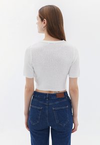 Bílý, krátký, cropovaný pletený top s texturovaným povrchem, spárovaný s modrými džíny s vysokým pasem, které mají koženou nášivku vzadu.