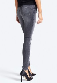 Persona vistiendo jeans ajustados negros deslavados y tacones altos negros con adornos de joyas, de pie contra un fondo blanco.