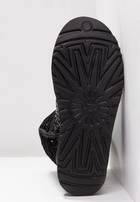 Semelle en caoutchouc noir d'une botte UGG montrant un motif de bande de roulement distinct et le logo, positionnée verticalement sur un fond blanc.