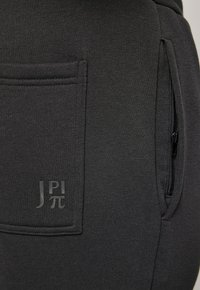 Donkergrijze joggers van zacht materiaal, voorzien van een zijvak met rits en een zichtbaar logo in een bijpassende donkere tint. Geweven afwerking.