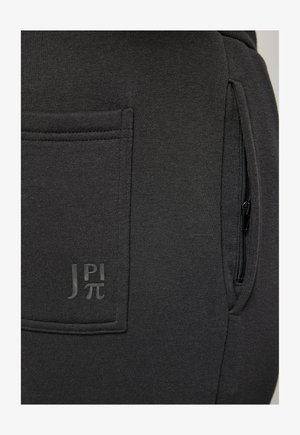 Tummanharmaat juoksuhousut, jotka on valmistettu pehmeästä kankaasta. Housuissa on sivuvertszip-pussi ja näkyvä logo samansävyisenä. Teksturoitu pinta.