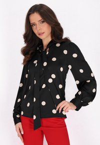 Blusa nera con grandi pois beige, con colletto annodato e maniche a sbuffo, abbinata a pantaloni rossi aderenti. Tessuto morbido.