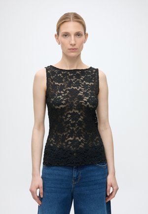 Femme à la peau claire et aux cheveux blonds porte un top sans manches en dentelle noire et un jean bleu, debout devant un fond clair uni.