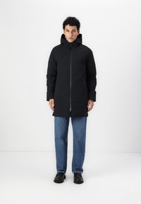 Svart vanntett parka med glidelås, hette og elastiske mansjetter. Båret over blå jeans og svarte sko, med et minimalistisk design.
