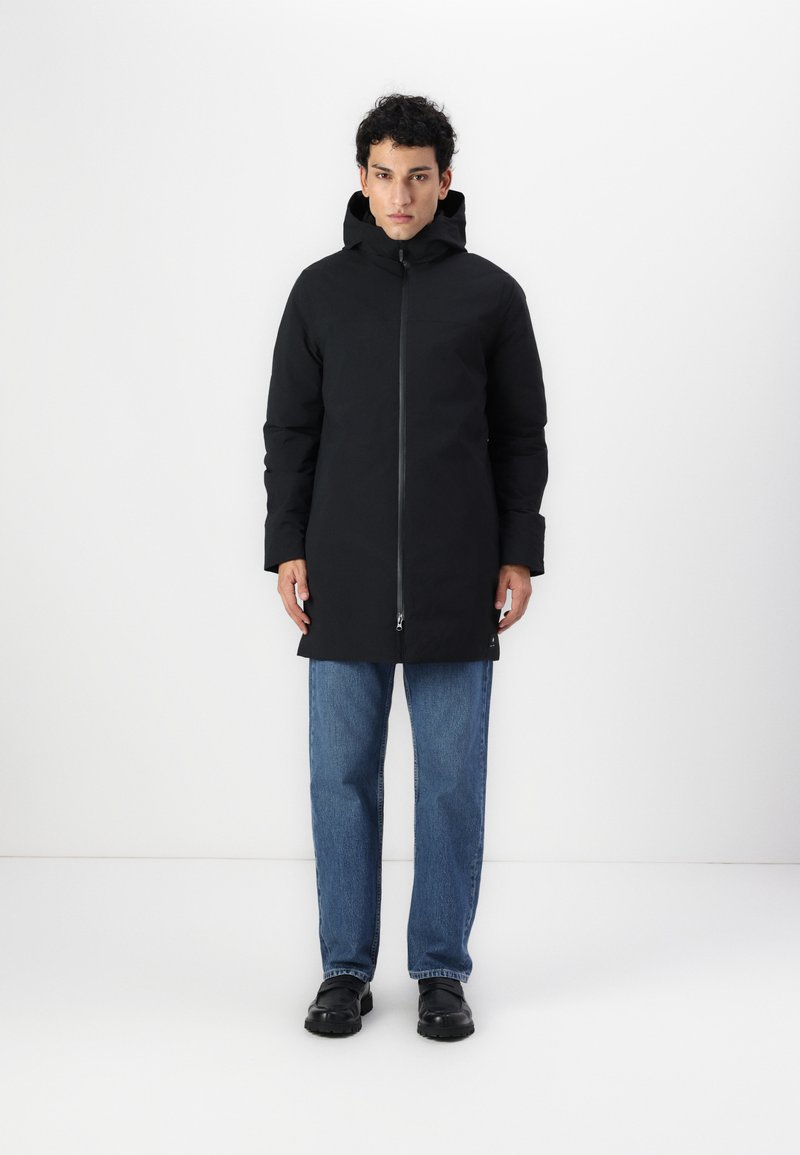 Svart vanntett parka med glidelås, hette og elastiske mansjetter. Båret over blå jeans og svarte sko, med et minimalistisk design.