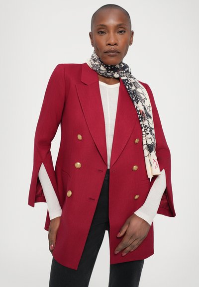 Red tailored blazer with double-breasted design, gold buttons, and open sleeve details. Worn over a white top with a patterned black and cream scarf.