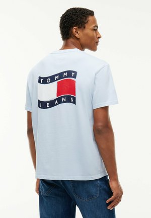 Uomo che indossa una maglietta azzurro chiaro con logo Tommy Jeans blu navy, bianco e rosso sulla schiena, abbinata a jeans di denim blu, visto di lato.