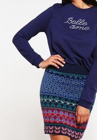 Donna che indossa un sweatshirt blu navy con la scritta "Belle âme" e una gonna di maglia colorata con disegni blu, verdi, rossi e rosa.