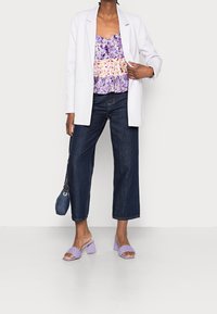 Donna che indossa un blazer bianco, una maglietta floreale viola, jeans cropped blu scuro, sandali con tacco lavanda e tiene in mano una borsa blu.