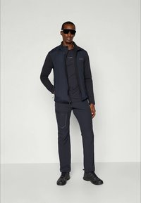 Un gilet sans manches bleu marine par-dessus un haut à manches longues noir, assorti à un pantalon. Présente une coupe slim et un design fonctionnel. Chaussures noires.