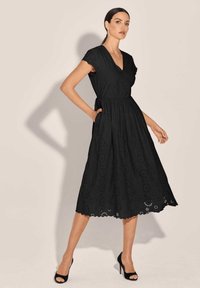 Schwarzes Kleid mit V-Ausschnitt, kurzen Ärmeln und einer ausgestellten A-Linie. Ein Ösenmuster und ein gewellter Saum verleihen Textur und Detail.