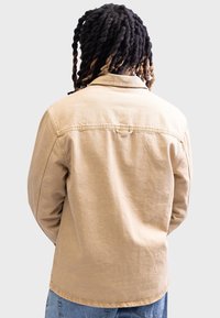 Chaqueta de mezclilla color beige con cuello, detalle de costura en el yugo trasero y un corte relajado. Costuras visibles y un tejido ligeramente texturizado.