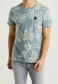 Lyhythihainen turkoosi t-paita, jossa on valkoisia kukkakuvioita ja pieni musta logopatchi vasemmassa rinnassa. Puuvillasekoitetta.