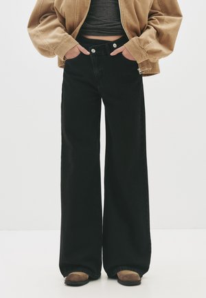 Jean large noir à taille haute, avec deux boutons en argent, des coutures discrètes et une finition soignée à l'ourlet. Associé à une veste en corduroy marron.