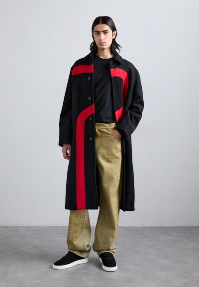 Cappotto di lana nero con motivo curvo rosso, camicia nera, pantaloni beige e sneakers neri. Figura isolata su uno sfondo neutro.
