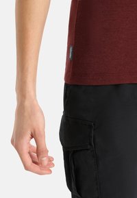 Burgundské tričko s krátkým rukávem spárované s černými cargo kalhotami, které mají boční kapsu a textilní etiketu na lemu trička.