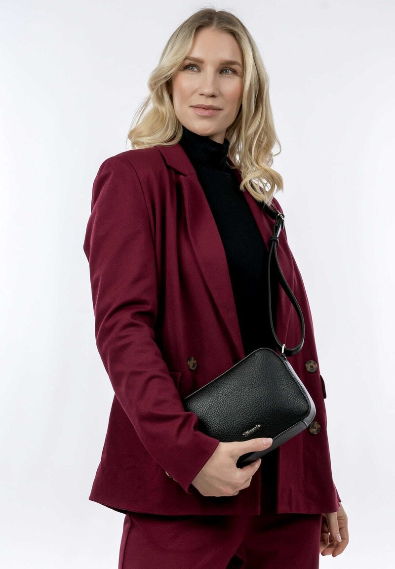 Black textured leather crossbody bag with adjustable strap, held in hand, paired with a burgundy suit jacket and black turtleneck.