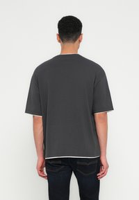 Man met kort krullend haar, gekleed in een losse donkergrijze t-shirt en donkere spijkerbroek, staande voor een effen witte muur.