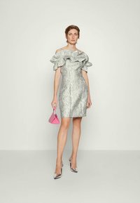 Ezüst fényű ruha fodros, vállat szabadon hagyó nyakkivágással, testhez simuló sziluettel és texturált mintával. A modell egy kis rózsaszín kézitáskát fog, ezüst magassarkú cipőben van.