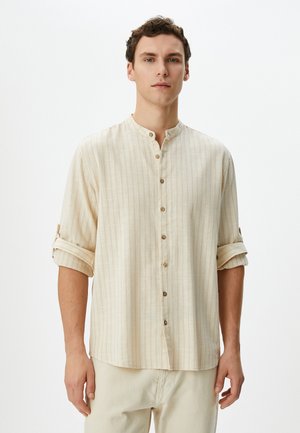 Camisa de botón beige claro con cuello tipo banda, rayas horizontales, mangas enrolladas y detalles de botones de madera. Tejido suave y texturizado.