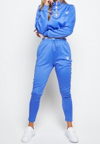 Blauer Sportanzug mit Reißverschlusskragen, enganliegende Hose und Kordelzugbund. Merkmale eines kontrastierenden Logos und Details mit elastischem Bund.