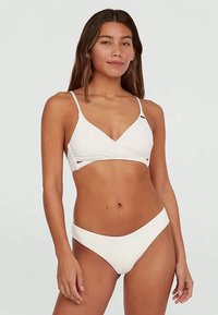 Žena v bílém bikinách s topem ve stylu wrap a vysokými kalhotkami, stojící na jednobarevném světle šedém pozadí.