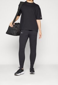 Černé tričko s logem, černé legíny s kapsami a černé sportovní boty. Osoba drží černou sportovní tašku s detaily z síťoviny.