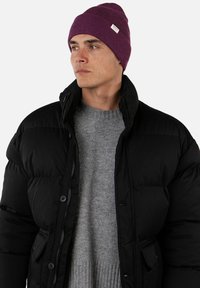 Black puffer jacket with a high collar, featuring a front zipper and button closures. Grey knitted sweater underneath and a purple beanie on top.