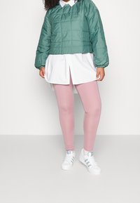 Grön quiltad pulloverjacka, vit lång skjorta under, ljusrosa leggings och vita sneakers med turkosa accenter. Släta texturer genom hela plagget.