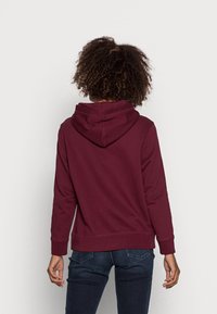 Burgundská mikina s kapucí vyrobená z měkké látky, s klokaní kapsou a žebrovanými manžetami. Zadní pohled ukazuje uvolněný střih a detail kapuce.