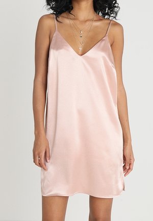 Vrouw draagt een mouwloze blush roze satijnen slipjurk met V-hals en gelaagde gouden kettingen tegen een effen achtergrond.